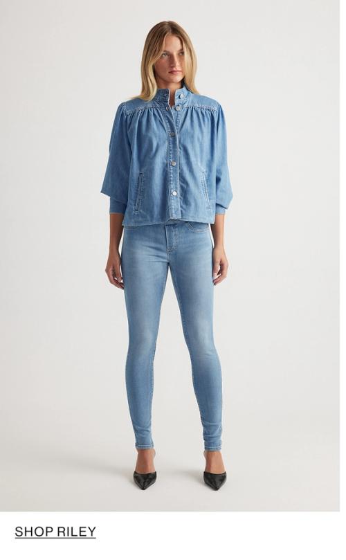 Woman standing in front of a white wall wearing a matching denim jacket and jeans set with texts that reads shop Riley