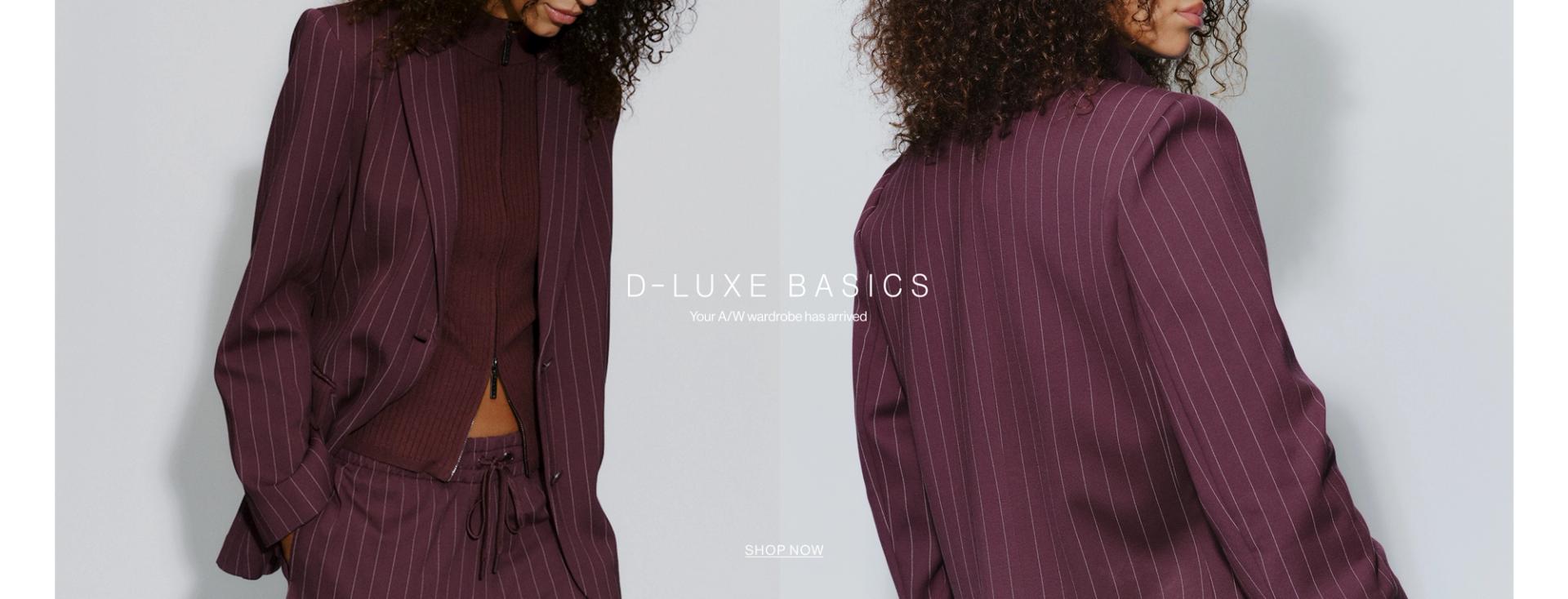 Model in a plum suit jacket standing in front of a white wall with text overlay which states "DLUXE BASICS"