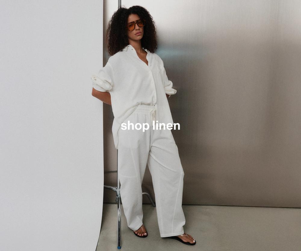Model in matching white linen shirt and long pants, standing casually against a metal wall with one hand behind her back. Text reads "Shop linen"