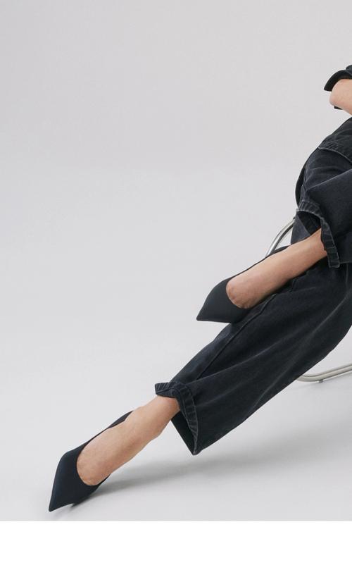 Woman sitting in a modern chair with her legs crossed and leaning to the side, wearing a black denim jacket and jeans set with black pointed heels.