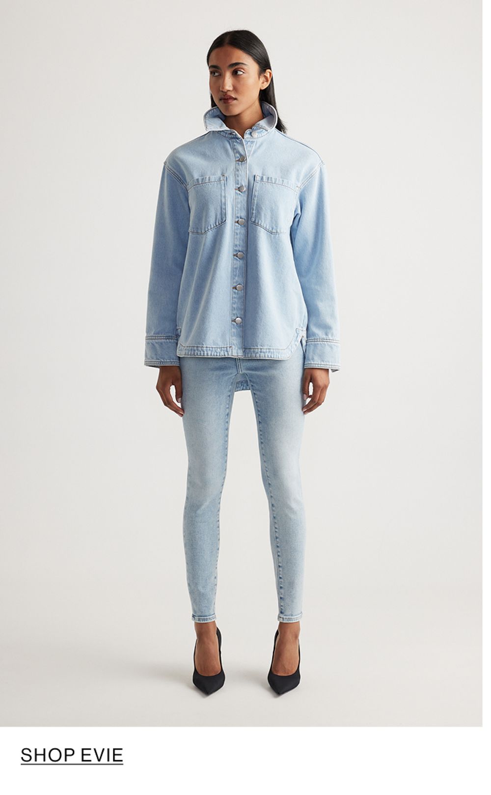 Woman standing in front of a white wall wearing a matching denim jacket and jeans set with texts that reads shop Evie
