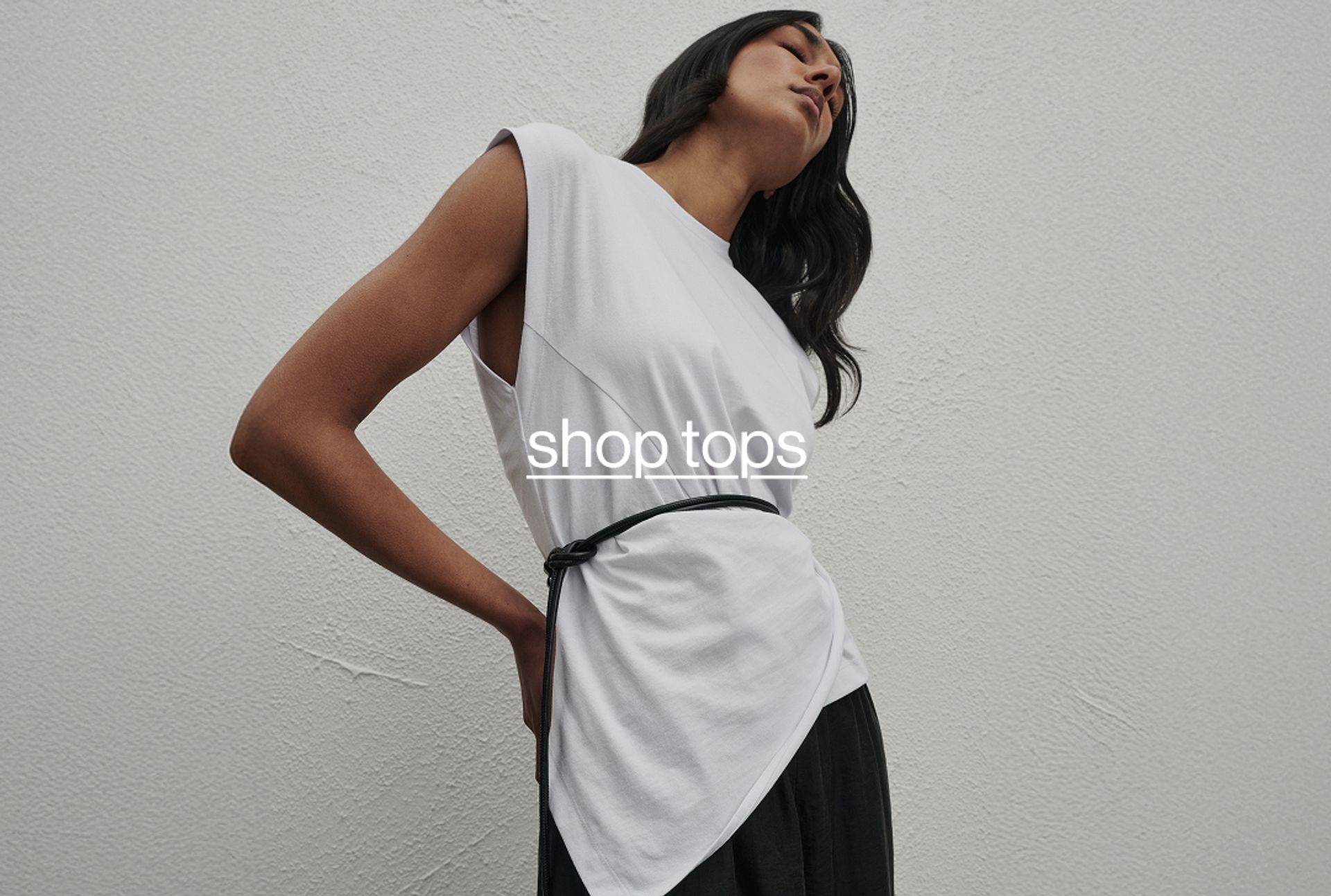 Woman standing in front of a white wall wearing a white top, with a think black belt. Text reads "shop tops"