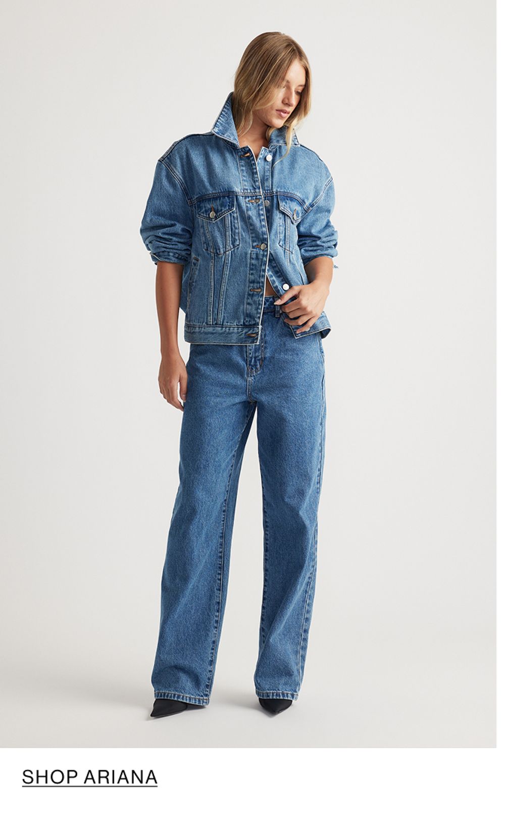 Woman standing in front of a white wall wearing a matching denim jacket and jeans set with texts that reads shop Ariana