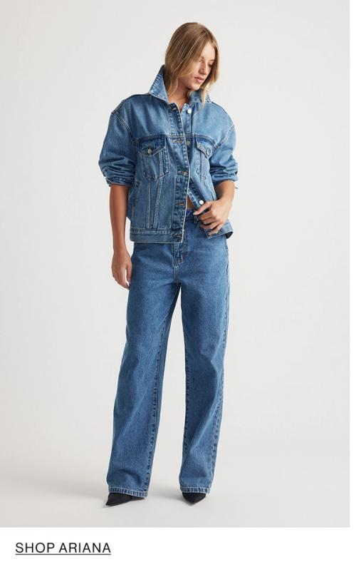 Woman standing in front of a white wall wearing a matching denim jacket and jeans set with texts that reads shop Ariana