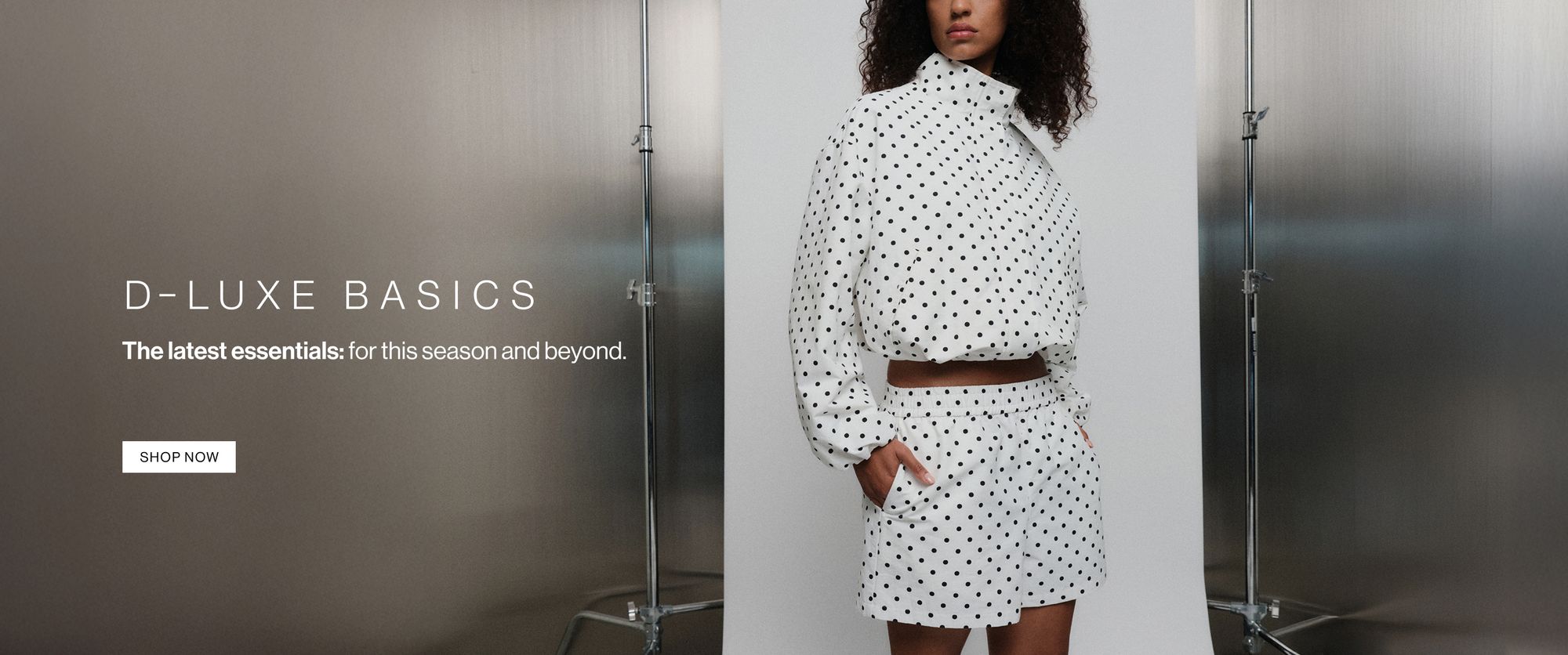 Model in a polka dot coordinated set, standing casually against a metal wall with one hand in her pocket. Text reads ‘DLUXE basics: the latest essentials for this season and beyond.