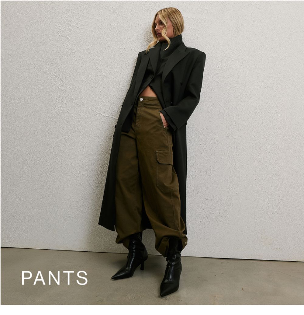 Woman wearing a long black coat, green pants and boots standing in front of a white wall with text that states 25% off pants
