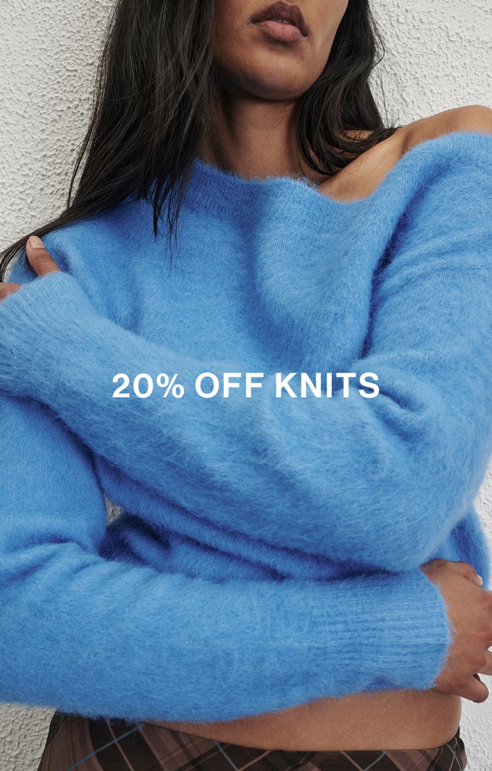 Woman in a blue knit in front of a white wall with text that reads "20% off all Knits"