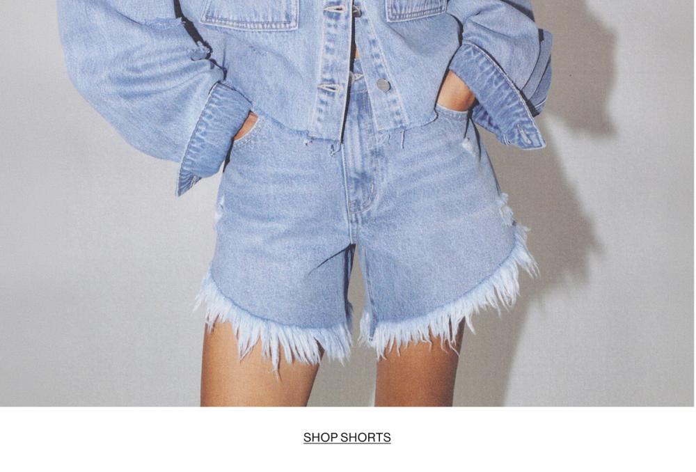 Woman posing with her hands in her pockets wearing denim shorts with text overlay "SHOP SHORTS"