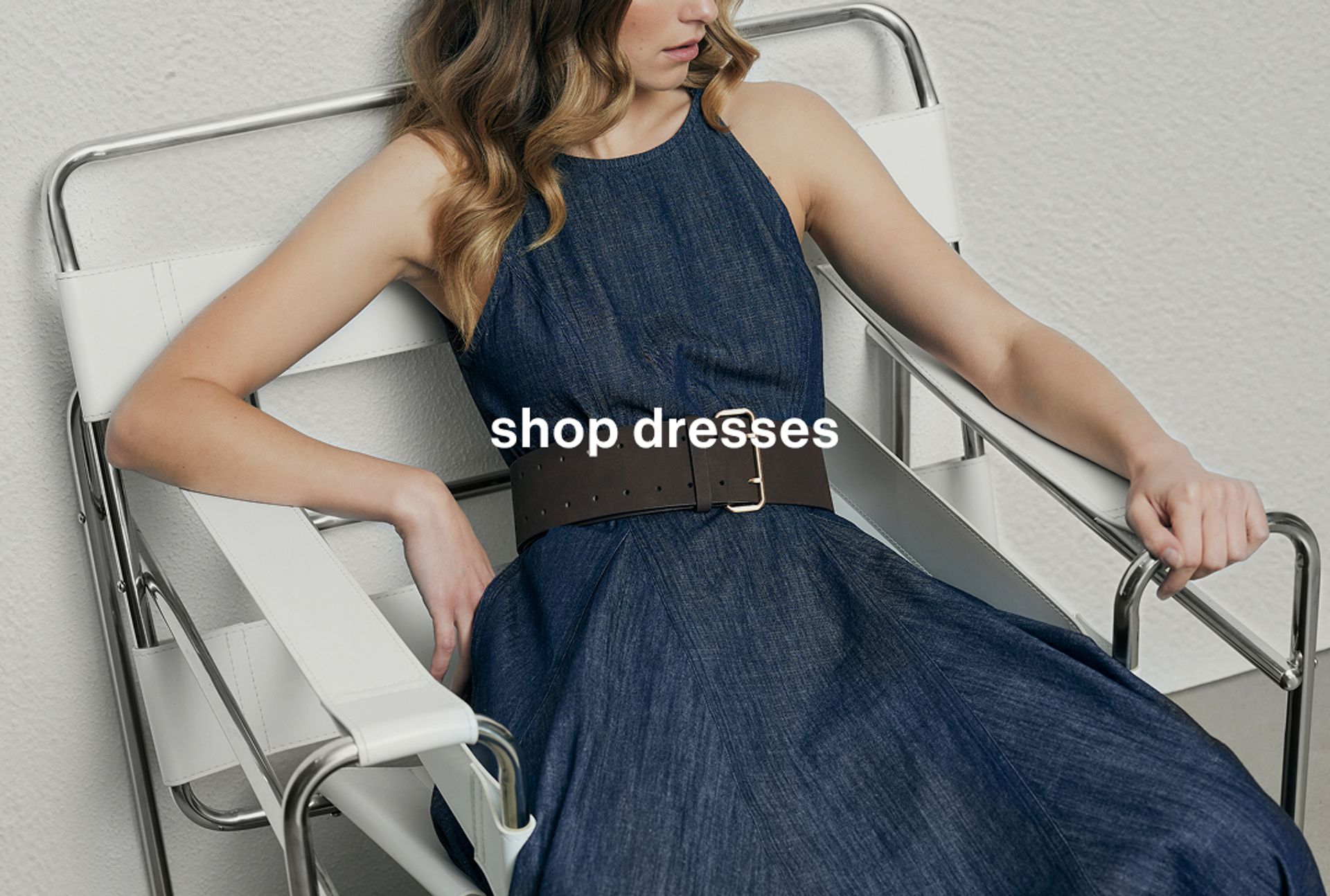 Woman wearing a blue long dress with a belt, seated in a modern chair. Text reads "shop dresses."
