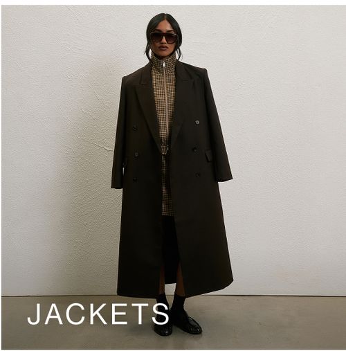 Woman posing in front of a white wall wearing a brown long coat with text that states "jackets"