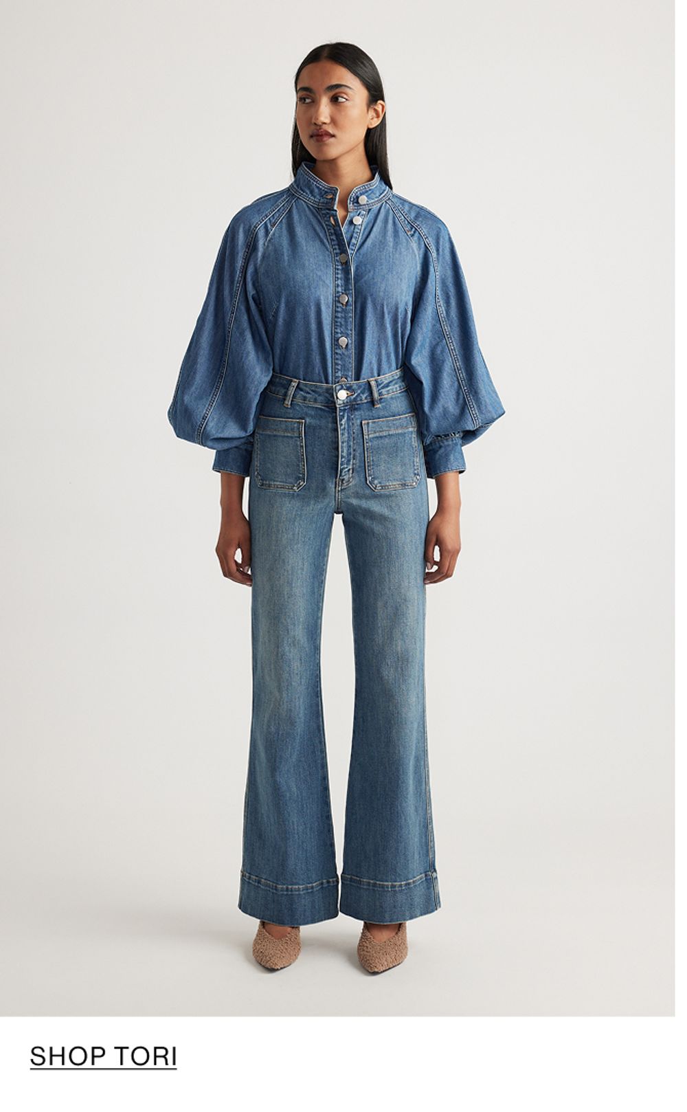 Woman standing in front of a white wall wearing a matching denim jacket and jeans set with texts that reads shop Tori