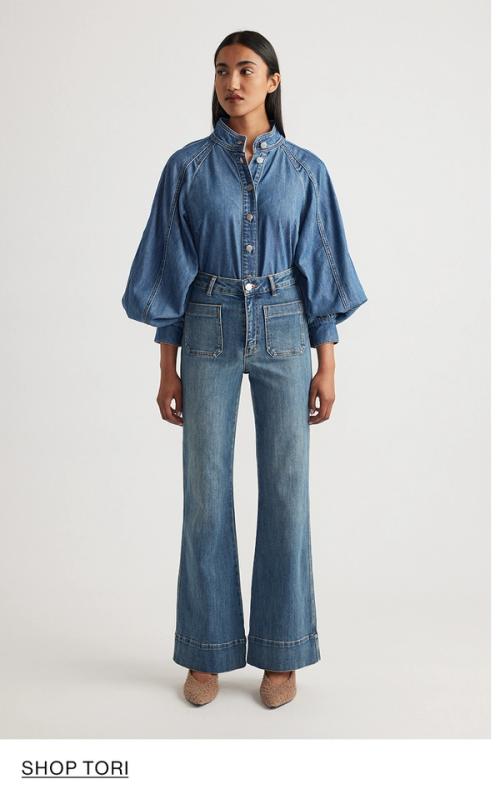 Woman standing in front of a white wall wearing a matching denim jacket and jeans set with texts that reads shop Tori