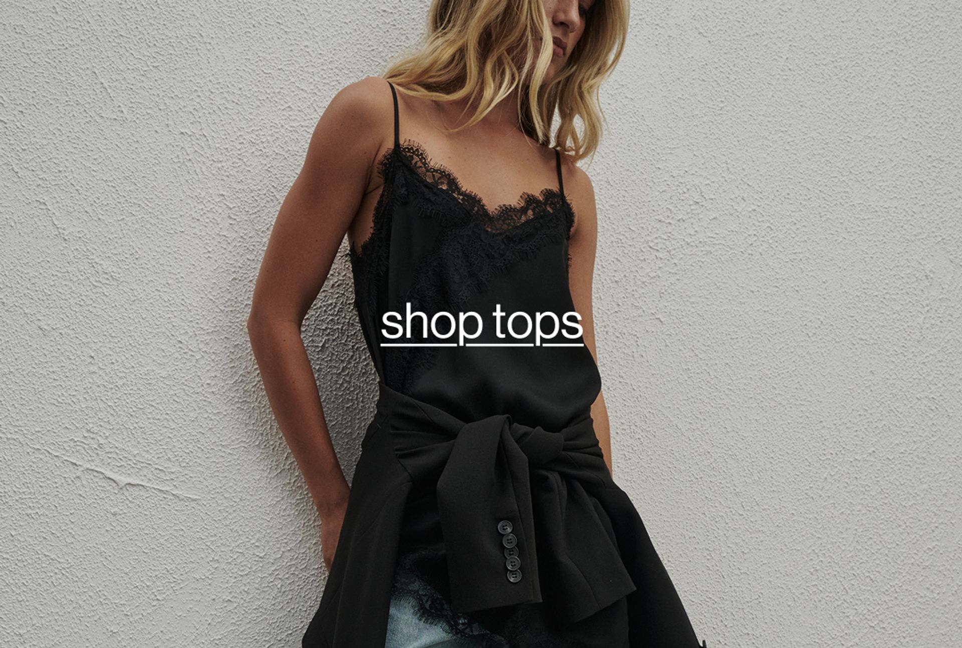 Woman standing in front of a white wall wearing a black lace top. Text reads "shop tops"