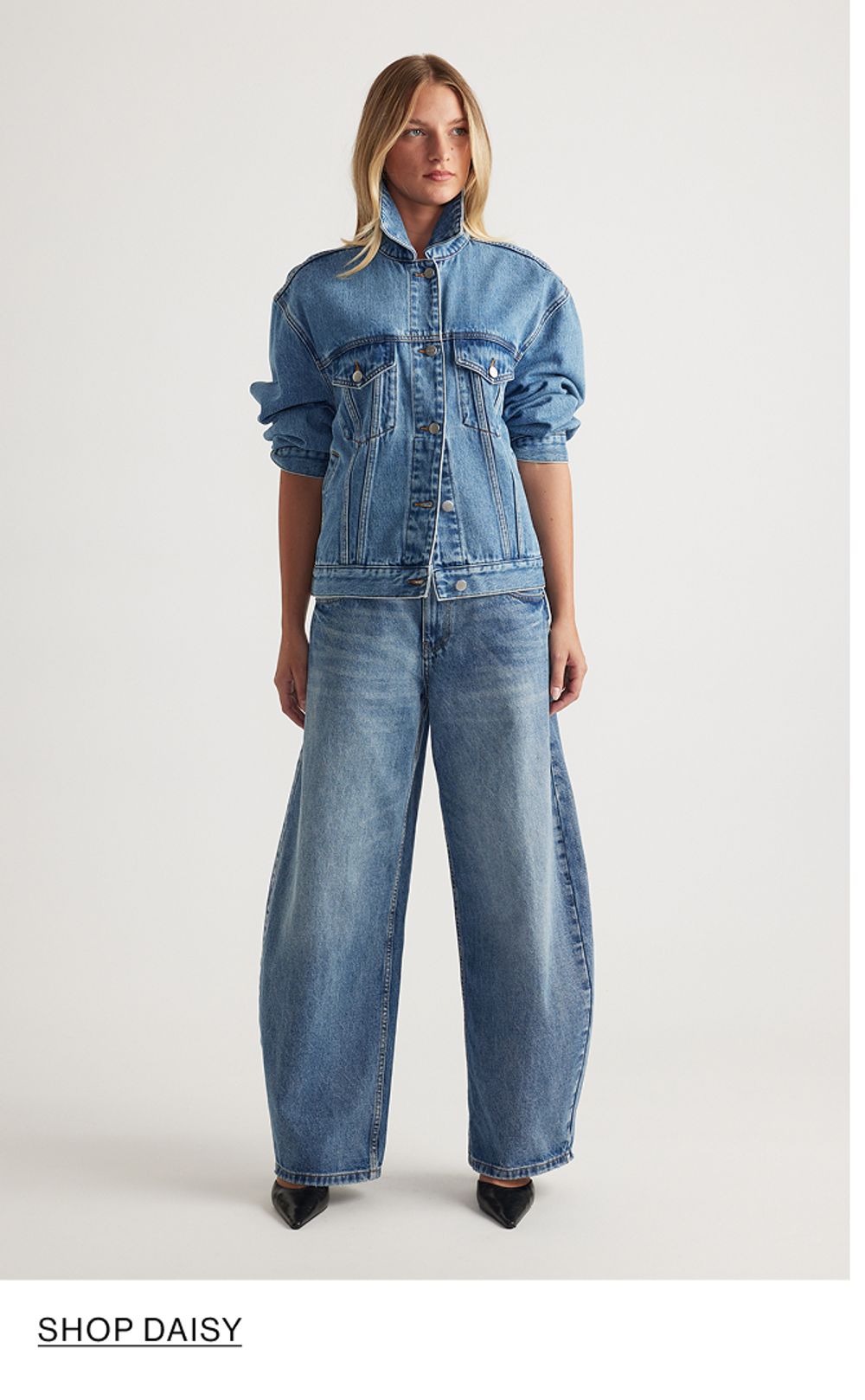 Woman standing in front of a white wall wearing a matching denim jacket and jeans set with texts that reads shop Daisy