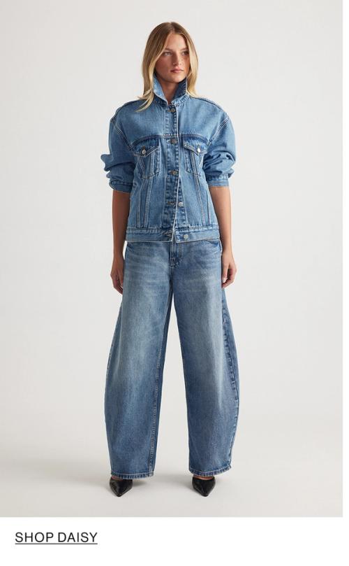 Woman standing in front of a white wall wearing a matching denim jacket and jeans set with texts that reads shop Daisy