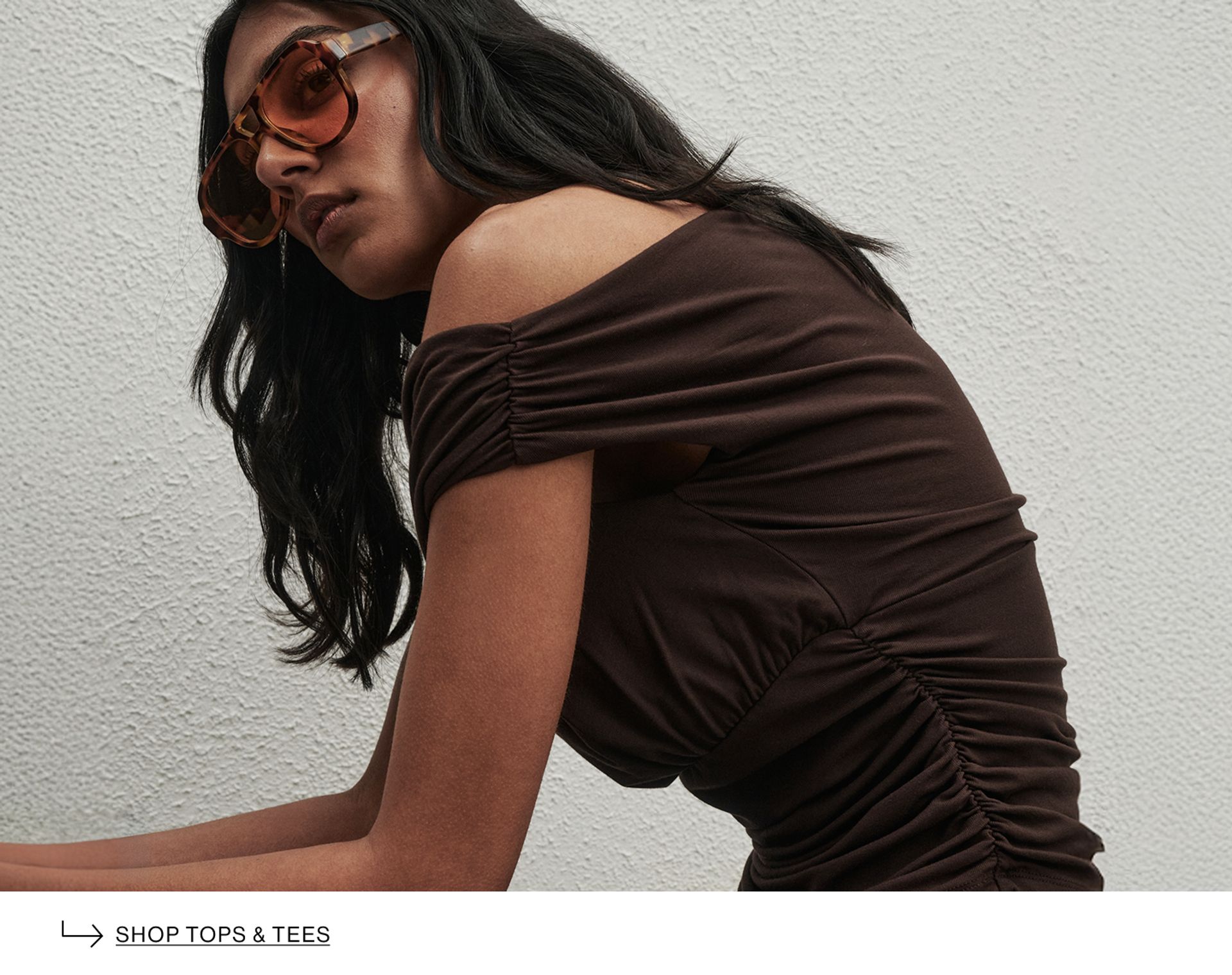 Woman wearing a brown top, and sunglasses. Text reads "shop tops & tees"