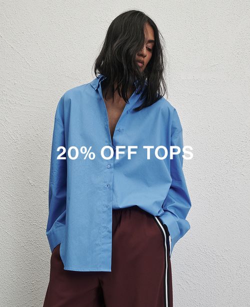 Woman standing in front of a white wall wearing a blue long sleeve shirt with text overlay which states "20% off Tops"