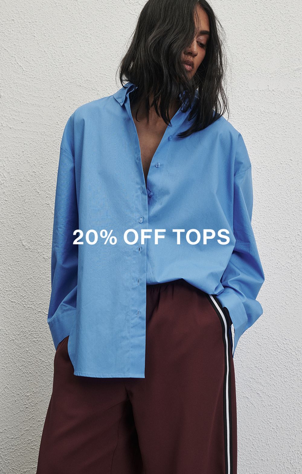 Woman standing in front of a white wall wearing a blue long sleeve shirt with text overlay which states "20% off Tops"
