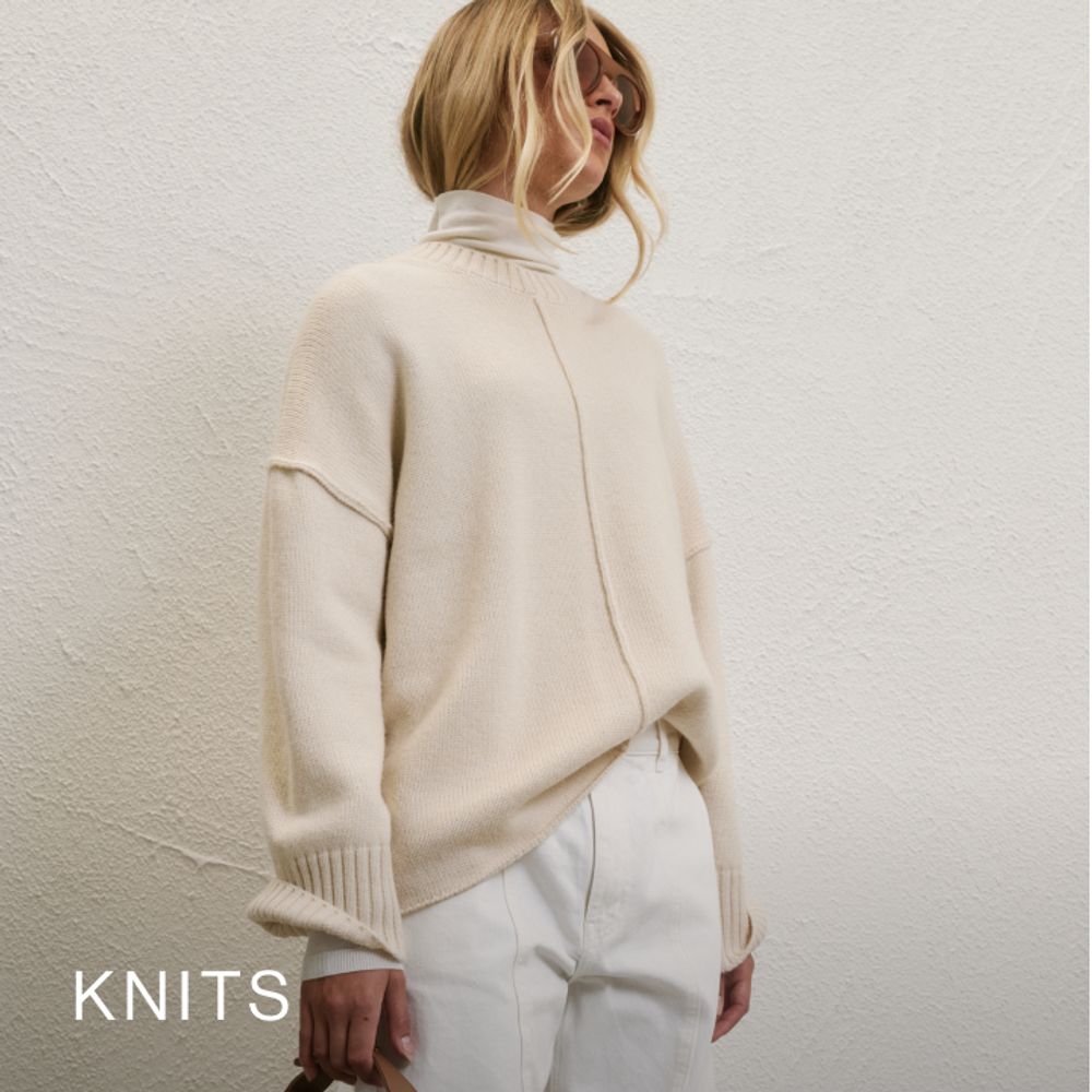 Woman posing in front of a white wall wearing a cream long sleeve knit with text that states "Knits"