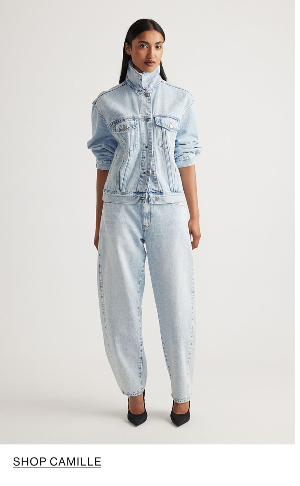 Woman standing in front of a white wall wearing a matching denim jacket and jeans set with texts that reads shop Camille