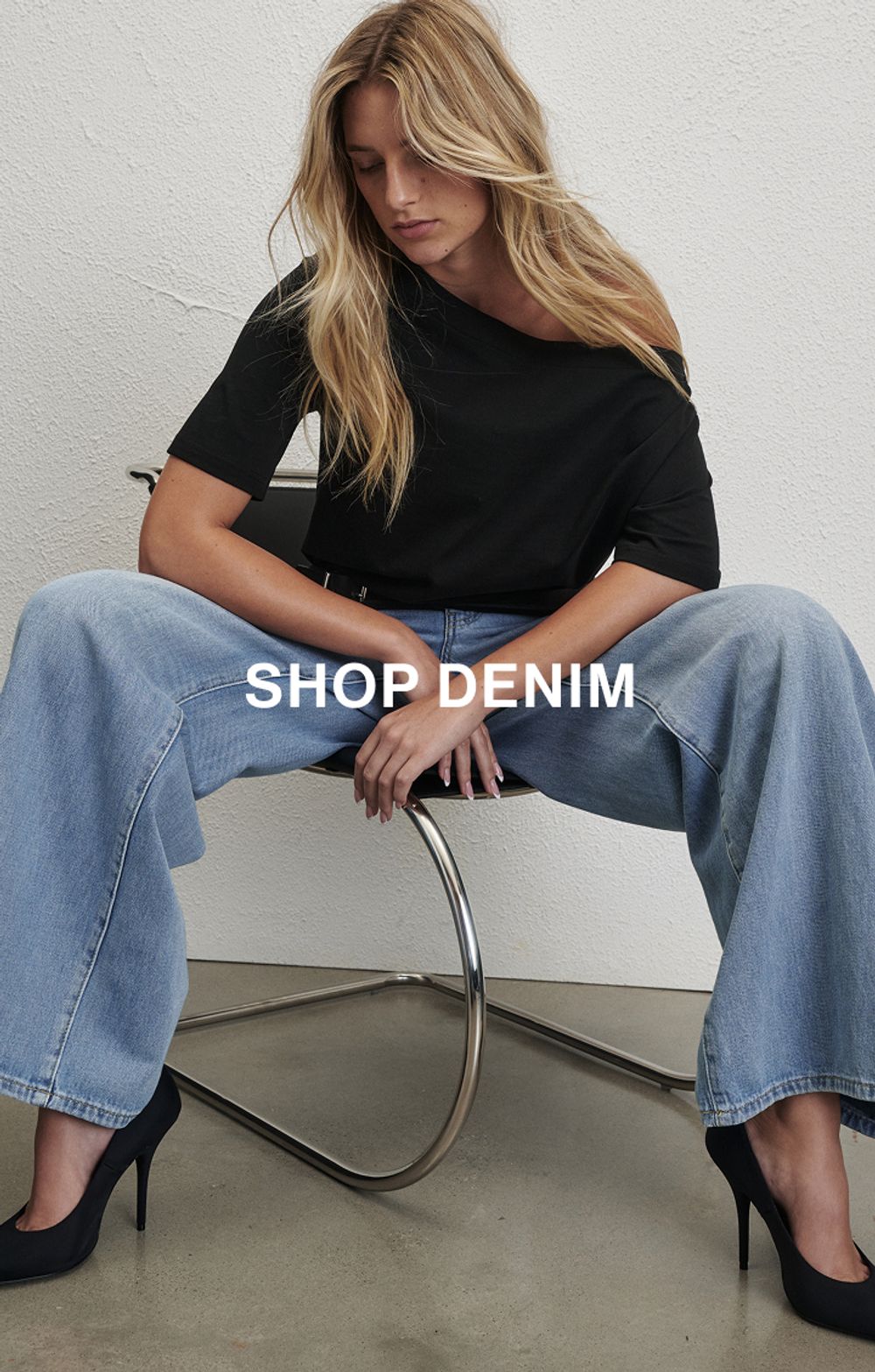 Woman posing in front of a white wall wearing a jeans and a black long sleeve top with text that states "20% off all denim"