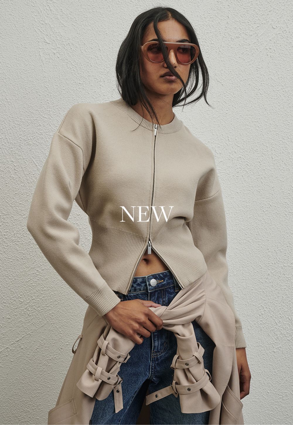 Woman standing in front of a white wall wearing a cream knit with text that reads "New"