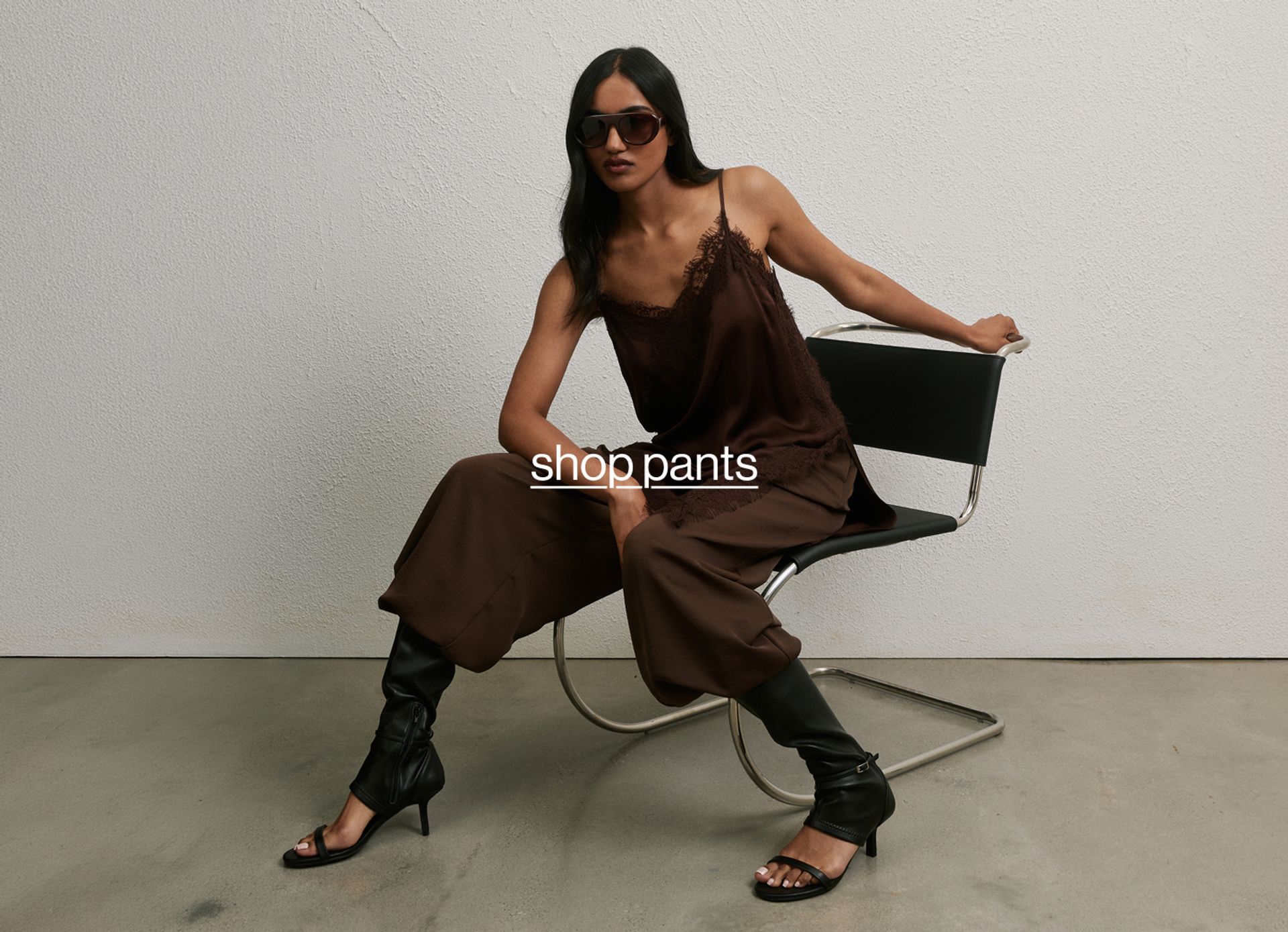 Woman posing wearing a black top and black long pants, standing in front of a white wall