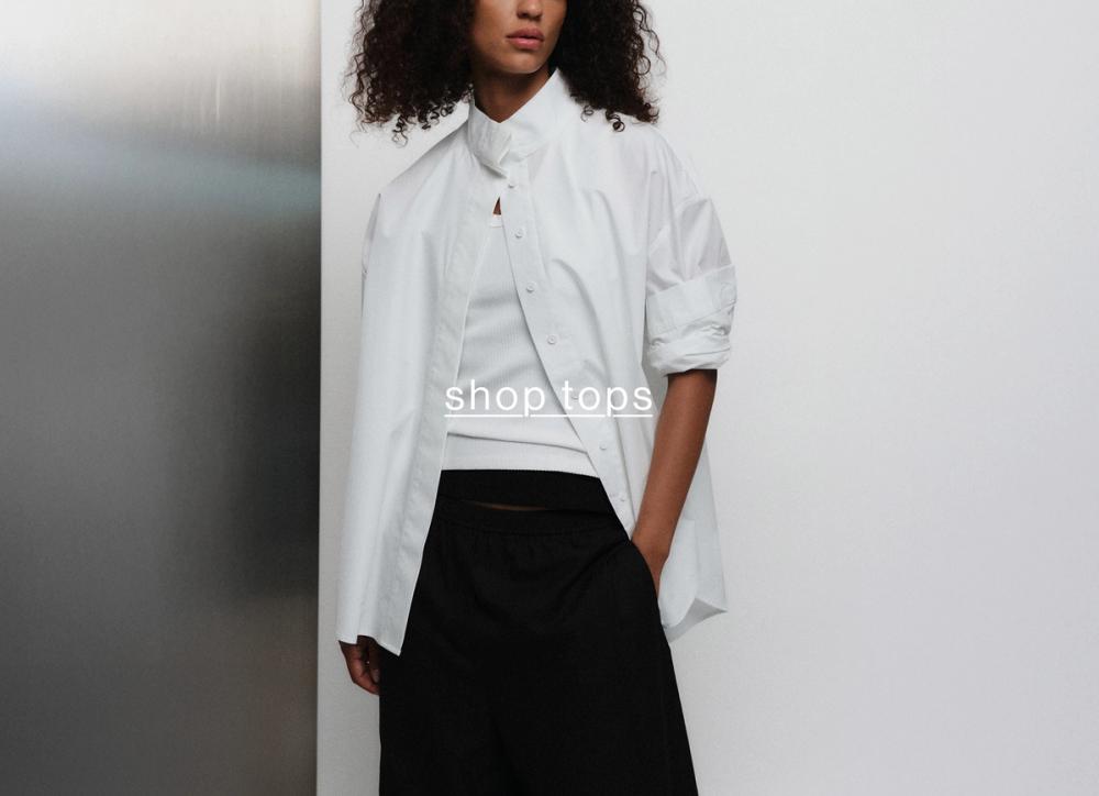 Model in white shirt, standing casually against a metal wall with her hand in her front pocket. Text reads "Shop tops"