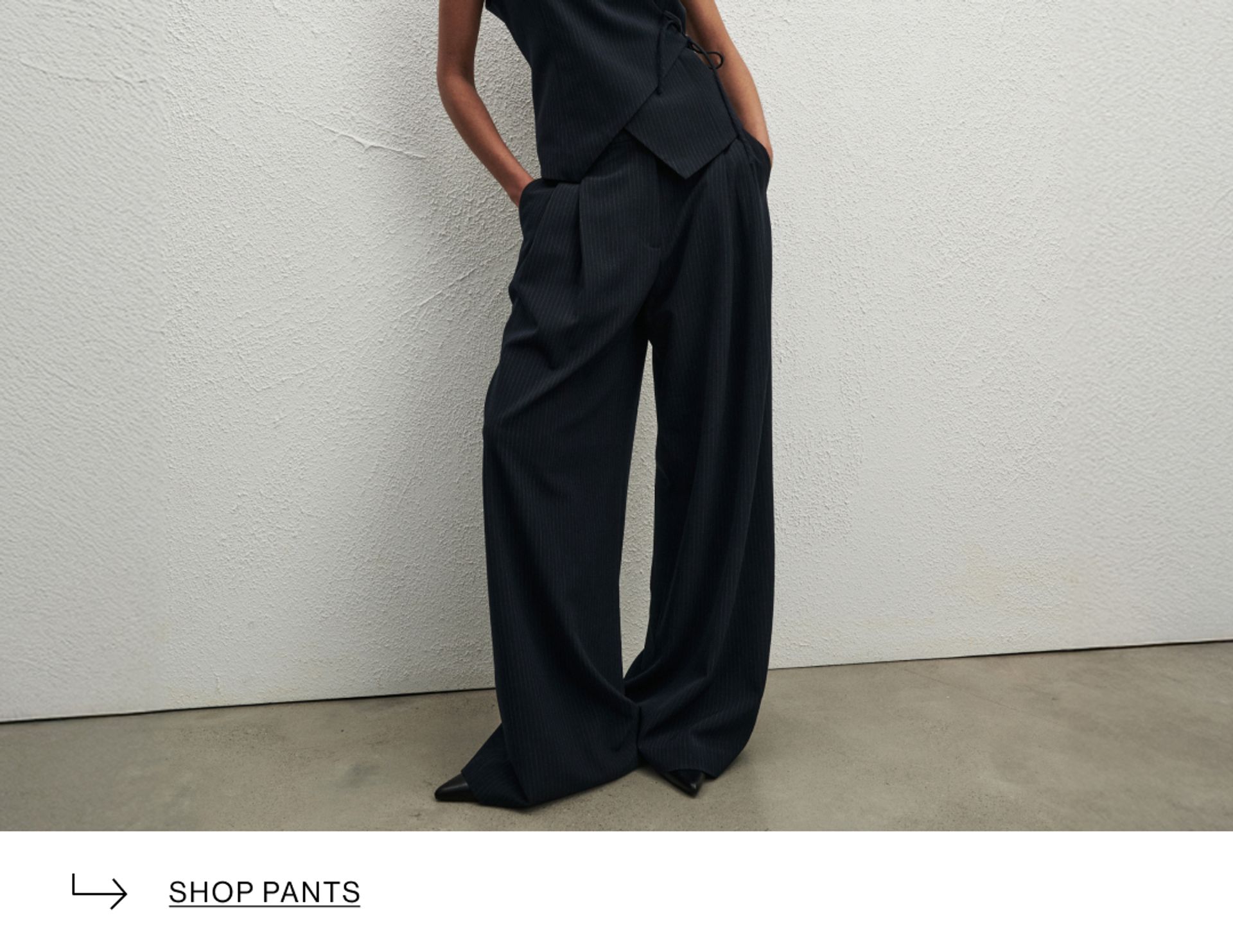 Woman posing wearing a black top and black long pants, standing in front of a white wall