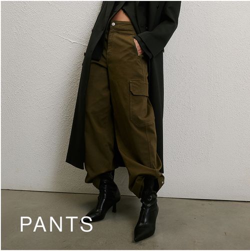 Woman wearing a long black coat, green pants and boots standing in front of a white wall with text that states 25% off pants