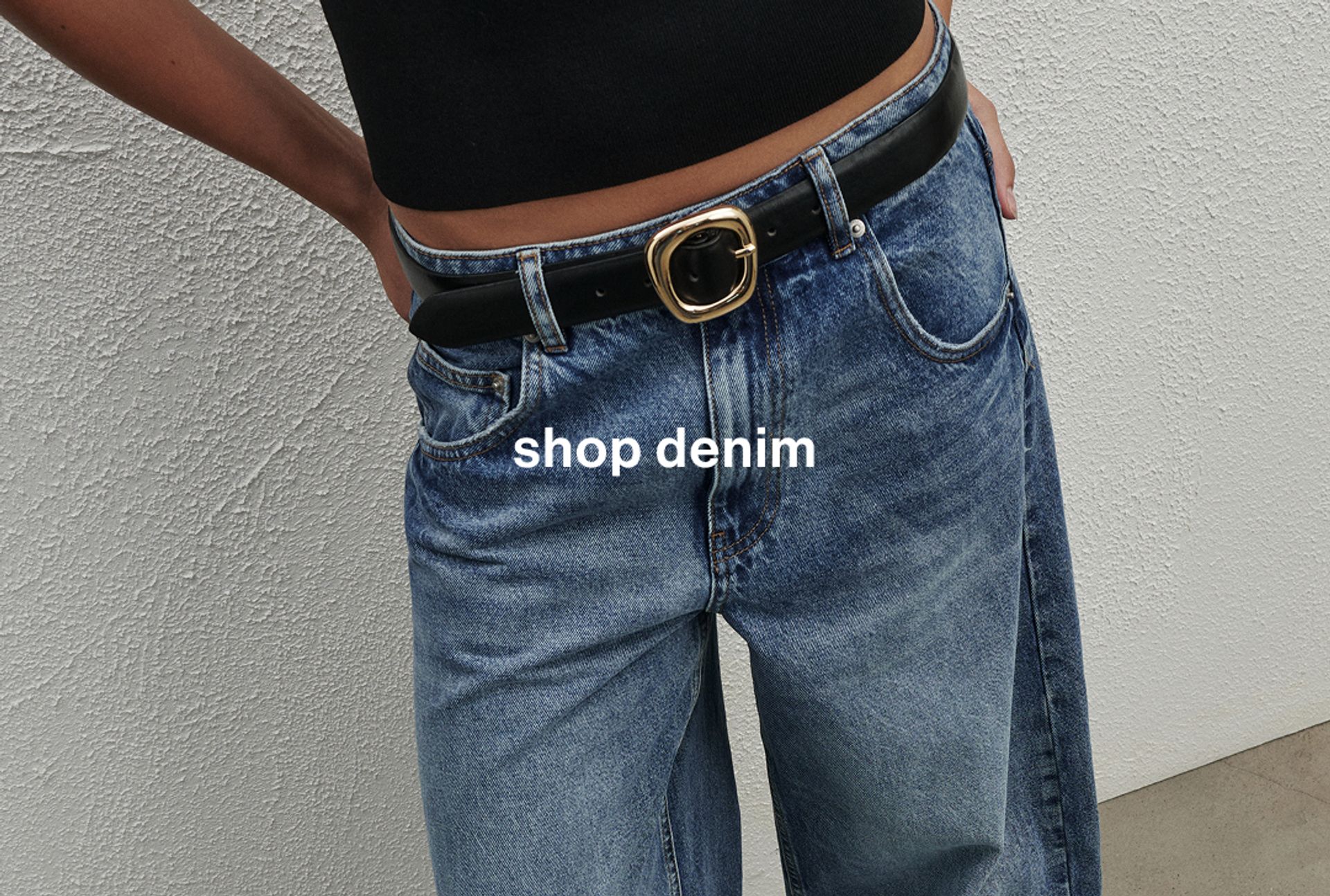 Woman wearing a black top, jeans, and a belt, standing in front of a white wall
