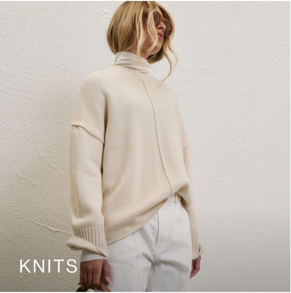 Woman posing in front of a white wall wearing a cream long sleeve knit with text that states "Knits"