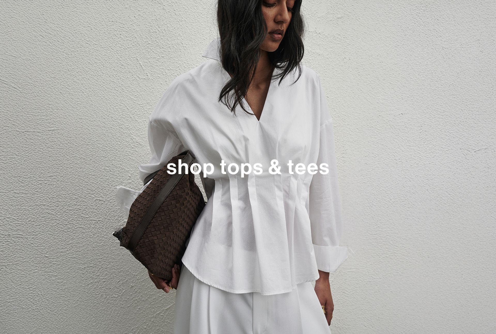 Woman standing in front of white wall with a white pleated shirt and collar, holding a black bag and staring at the ground.