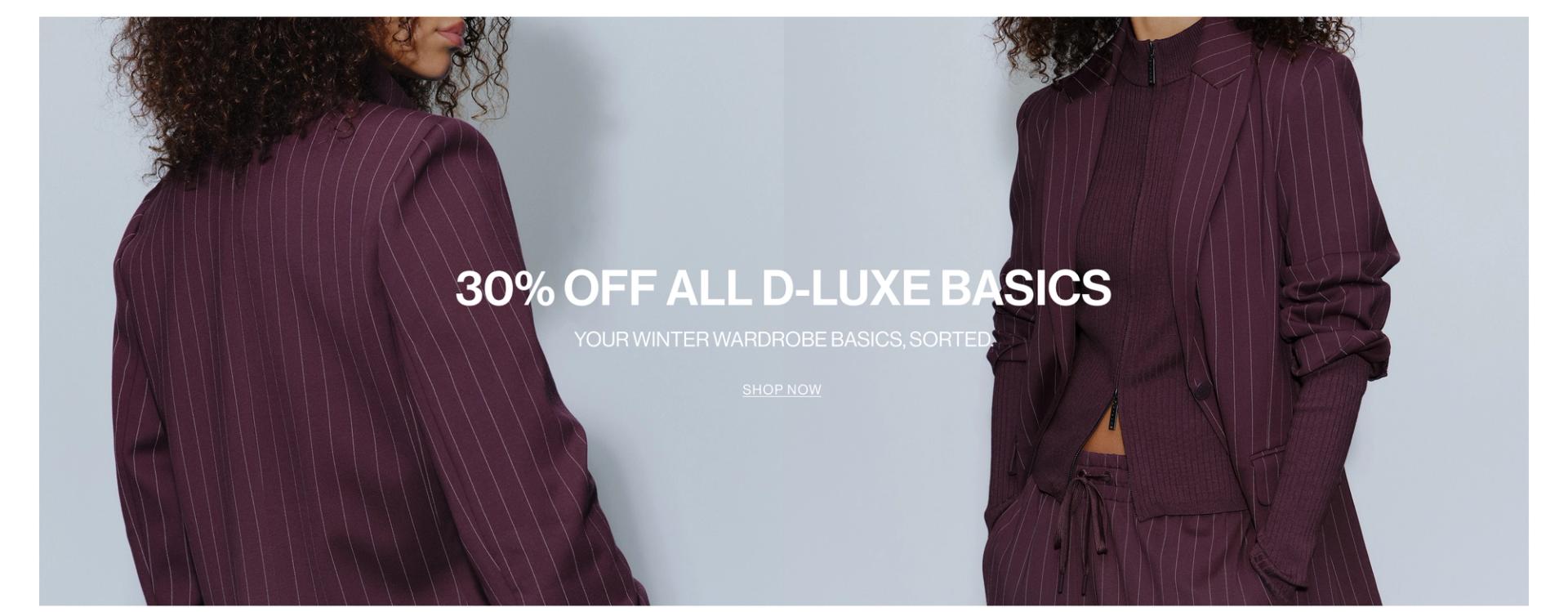 Model in a plum suit jacket standing in front of a white wall with text overlay which states "DLUXE BASICS"