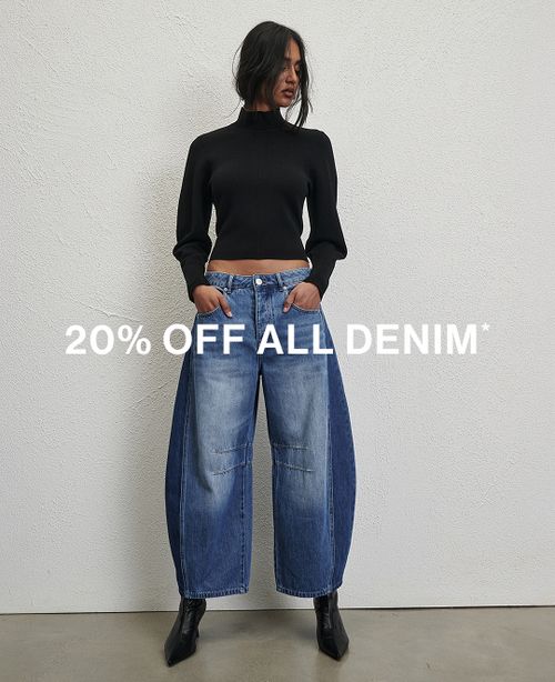Woman posing in front of a white wall wearing a jeans and a black long sleeve top with text that states "20% off all denim"