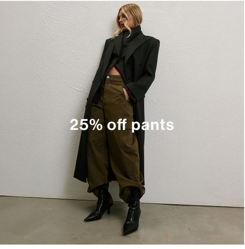 Woman wearing a long black coat, green pants and boots standing in front of a white wall with text that states 25% off pants