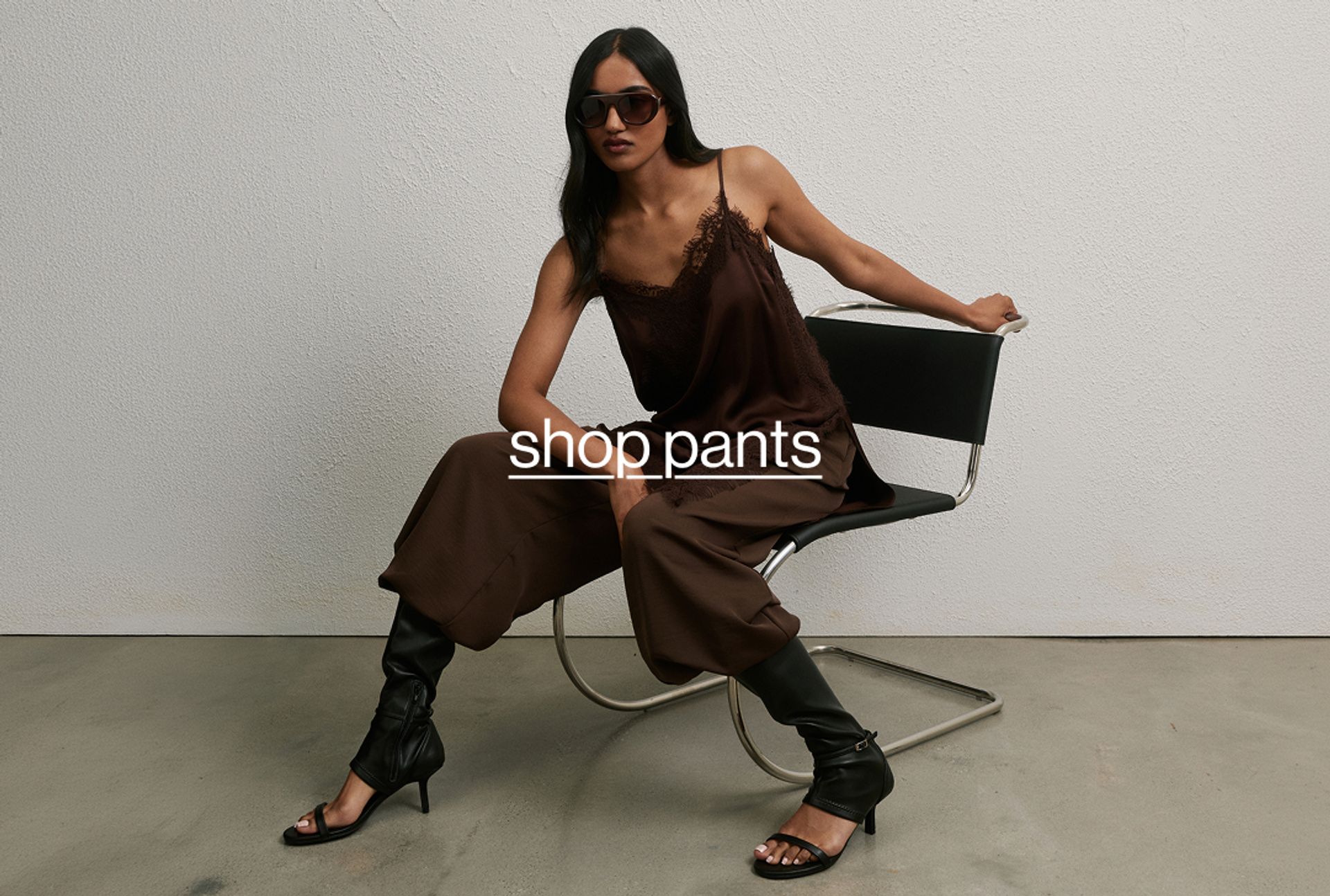 Woman posing wearing a black top and black long pants, standing in front of a white wall