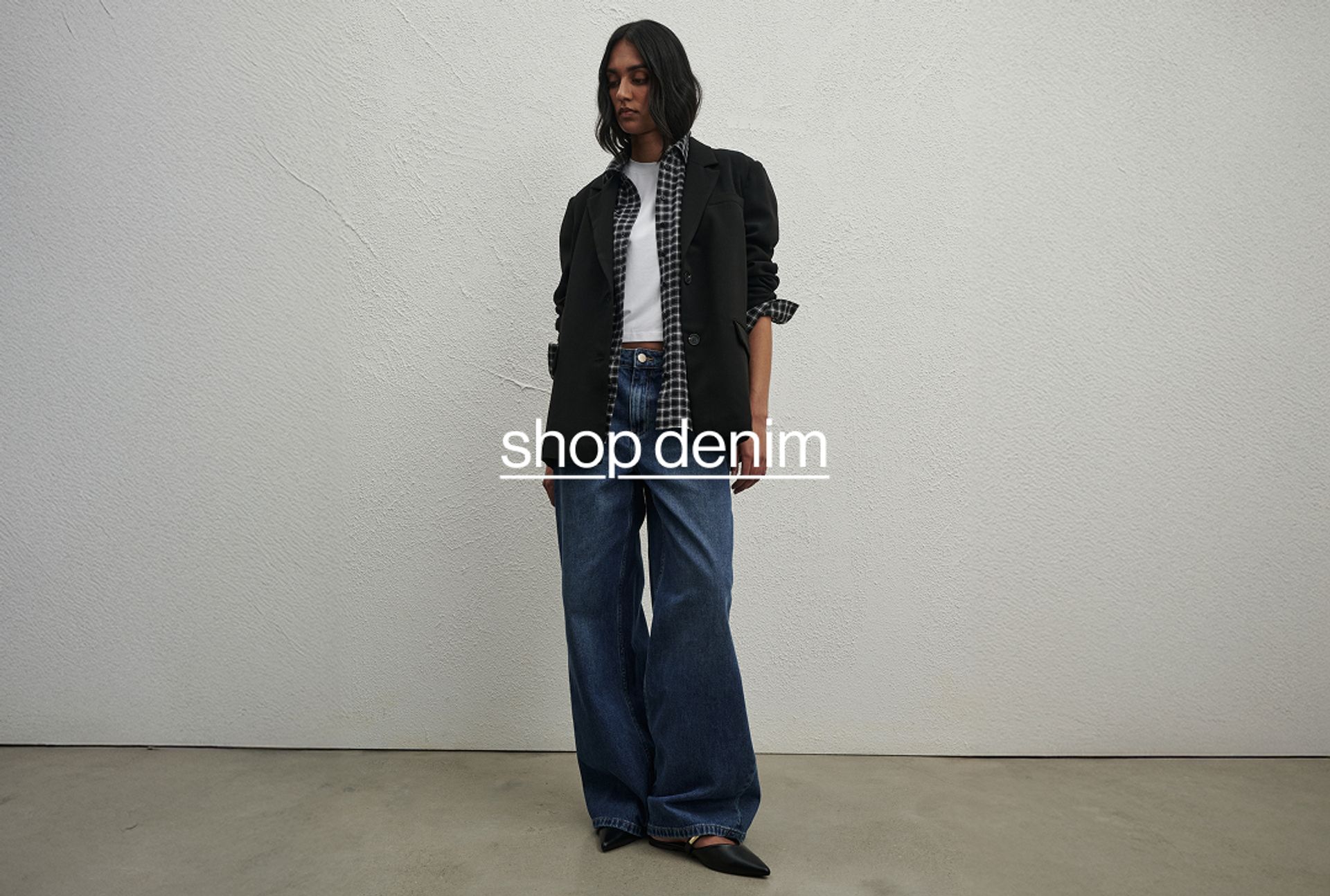 Model wearing on a black jacket, wearing denim jeans, standing against a minimalist white wall and gazing downward.
