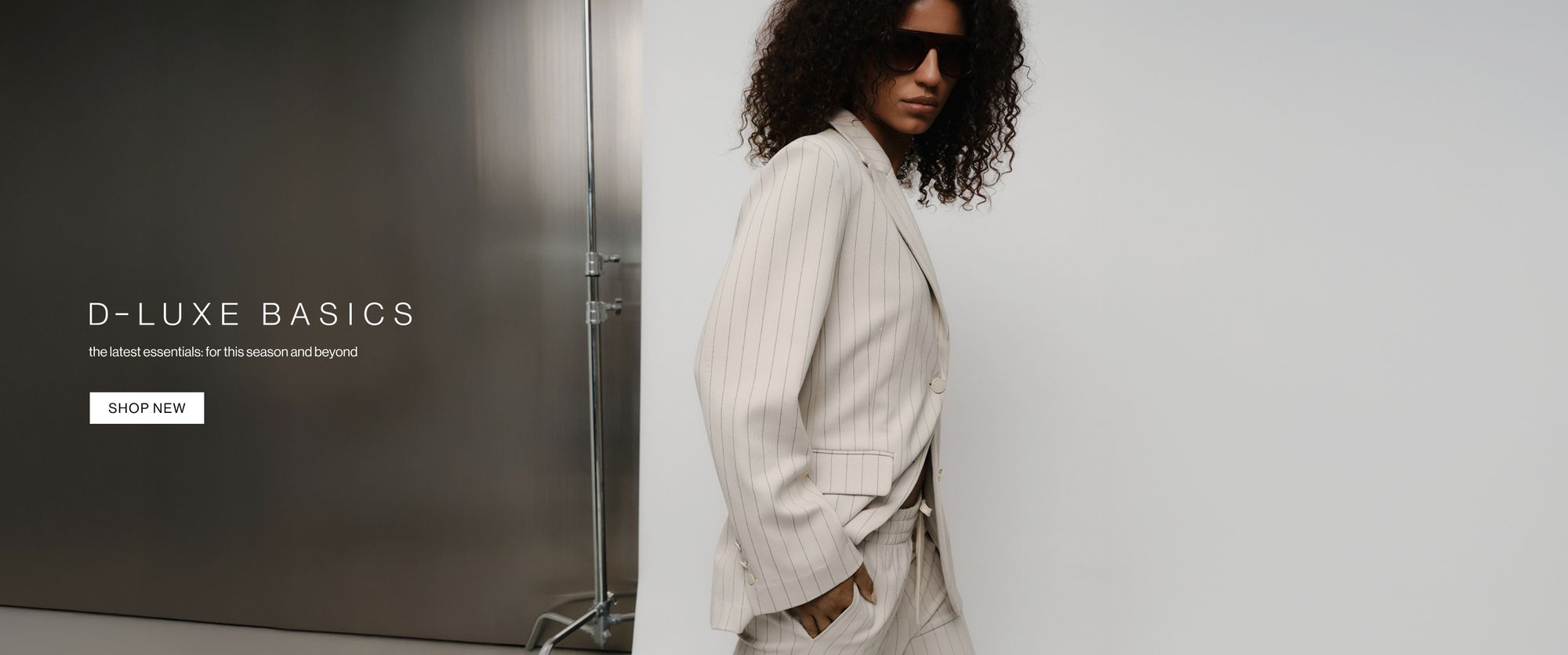 Model in a neutral coloured pinstripe suit posing against a metal wall with one hand in her pocket. Text reads ‘DLUXE basics: the latest essentials for this season and beyond.