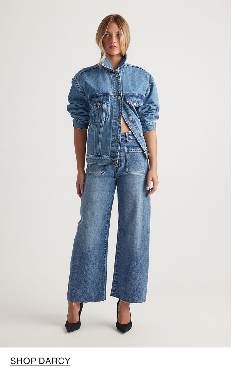 Woman standing in front of a white wall wearing a matching denim jacket and jeans set, with overlaid text that reads “Shop Darcy.”