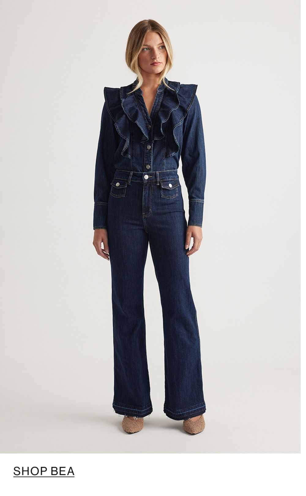 Woman standing in front of a white wall wearing a matching denim jacket and jeans set with texts that reads shop Bea