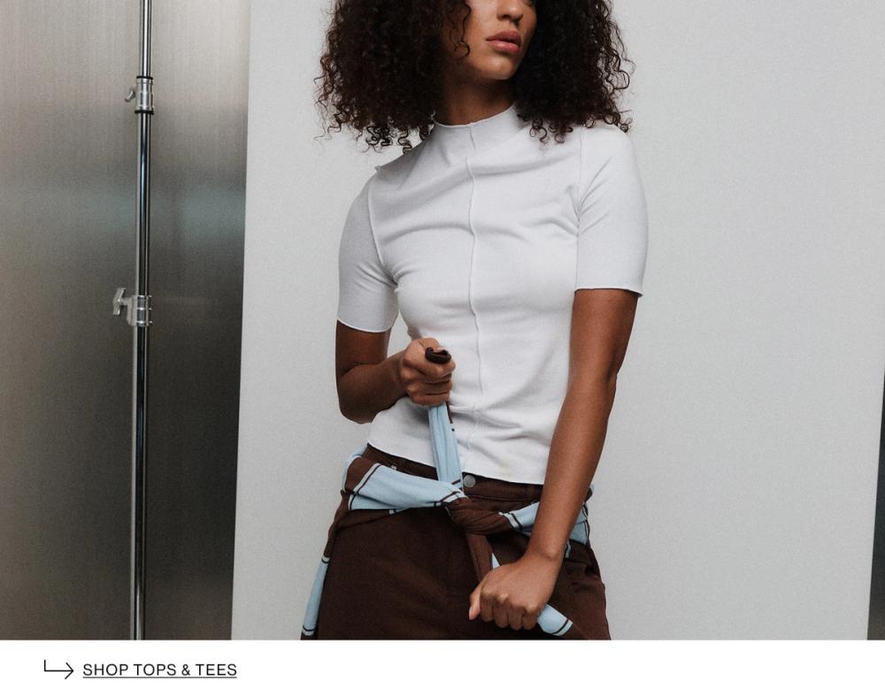 Model in white t-shirt, standing casually against a metal wall with her hand crossing a jumper around her waist. Text reads "Shop tops & tees"