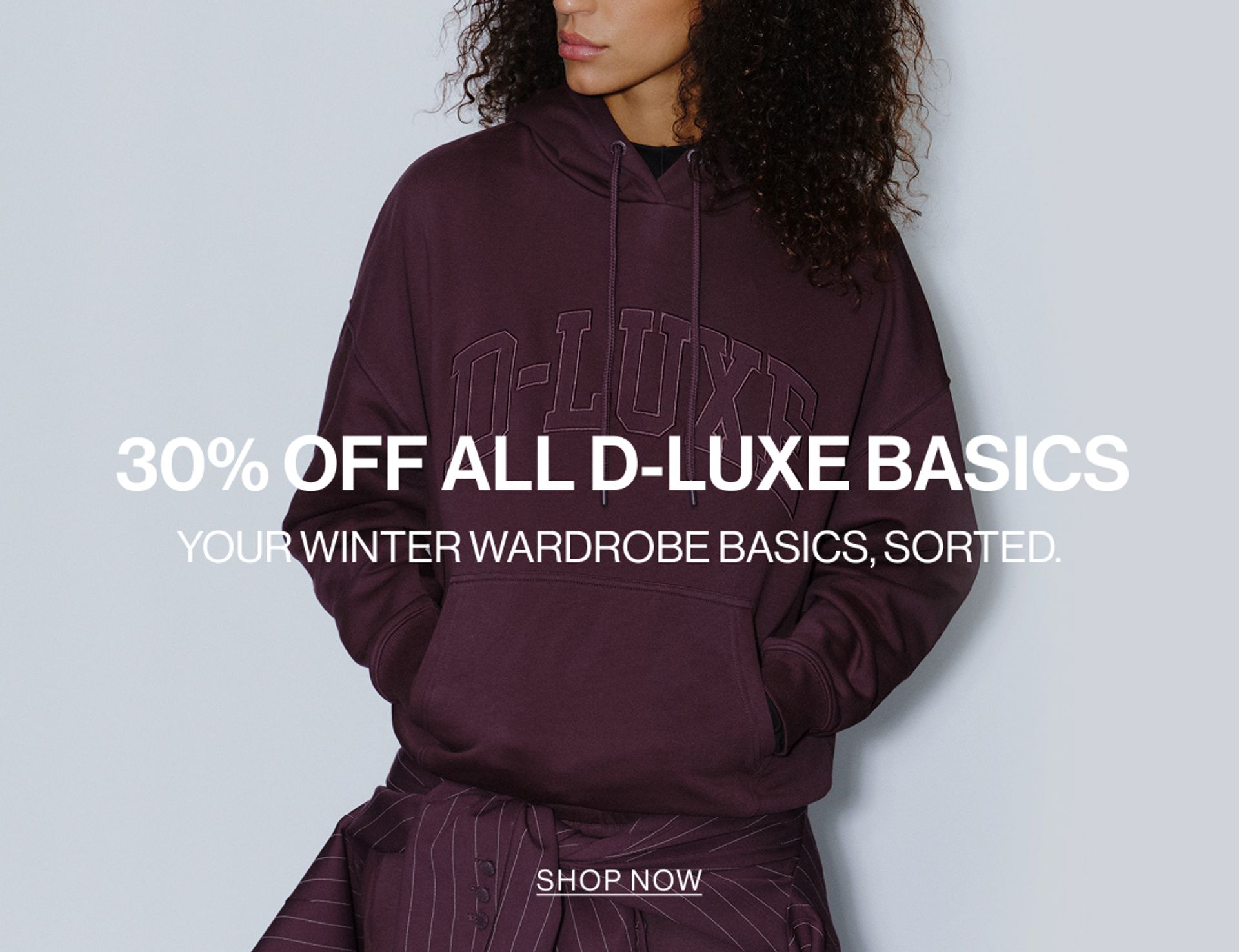 Model in a plum hoodie standing in front of a white wall with text overlay which states "DLUXE BASICS"