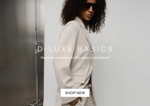 Model in a neutral coloured pinstripe suit posing against a metal wall with one hand in her pocket. Text reads ‘DLUXE basics: the latest essentials for this season and beyond.