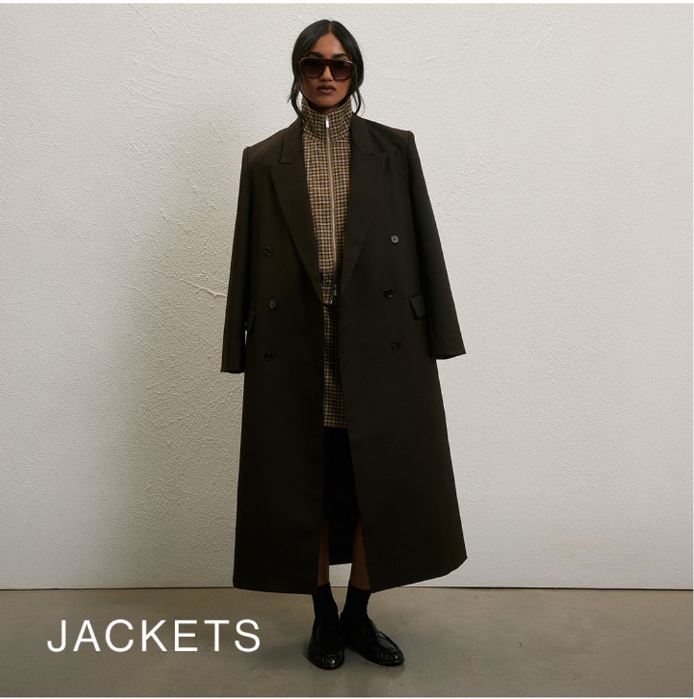 Woman posing in front of a white wall wearing a brown long coat with text that states "jackets"