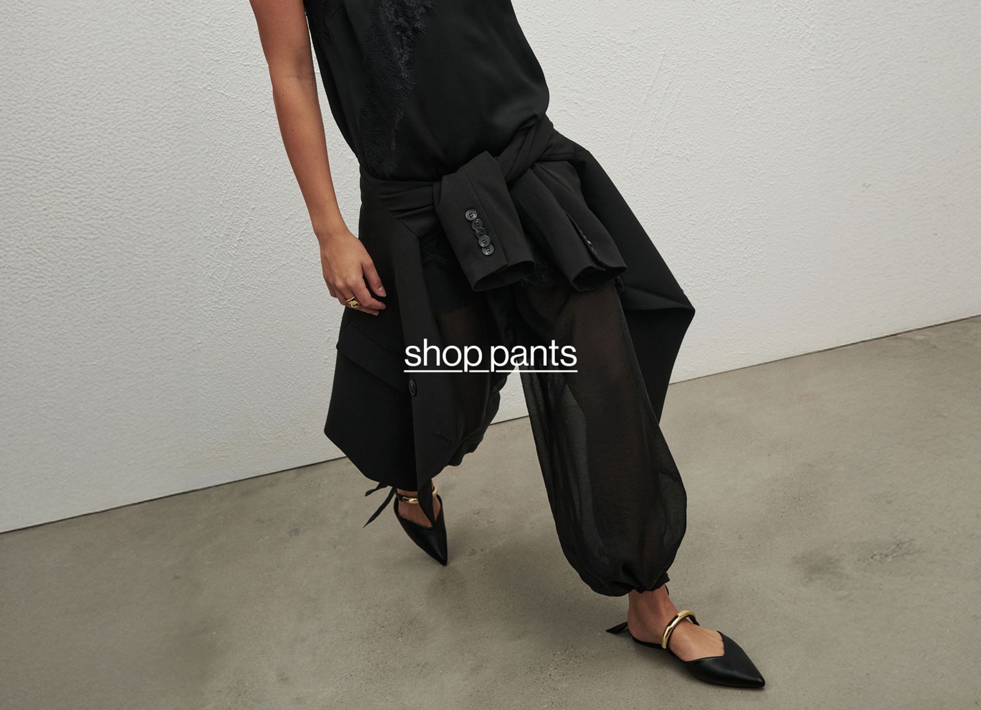 Woman posing wearing a black top and black long pants, standing in front of a white wall