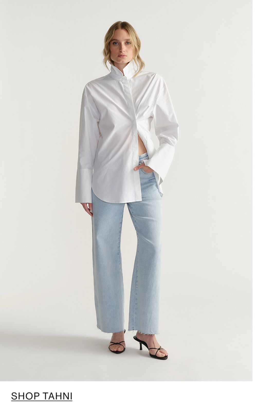 Woman standing in front of a white wall wearing a white shirt and light denim jeans, with overlaid text that reads “Shop Paloma”