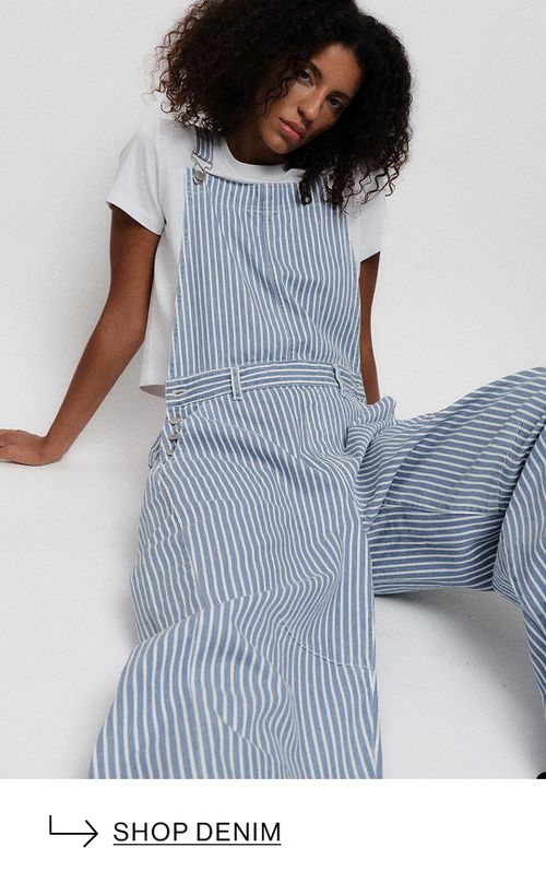 Model wearing a blue striped overalls, sitting in front of a white wall. Text reads ‘Shop denim.