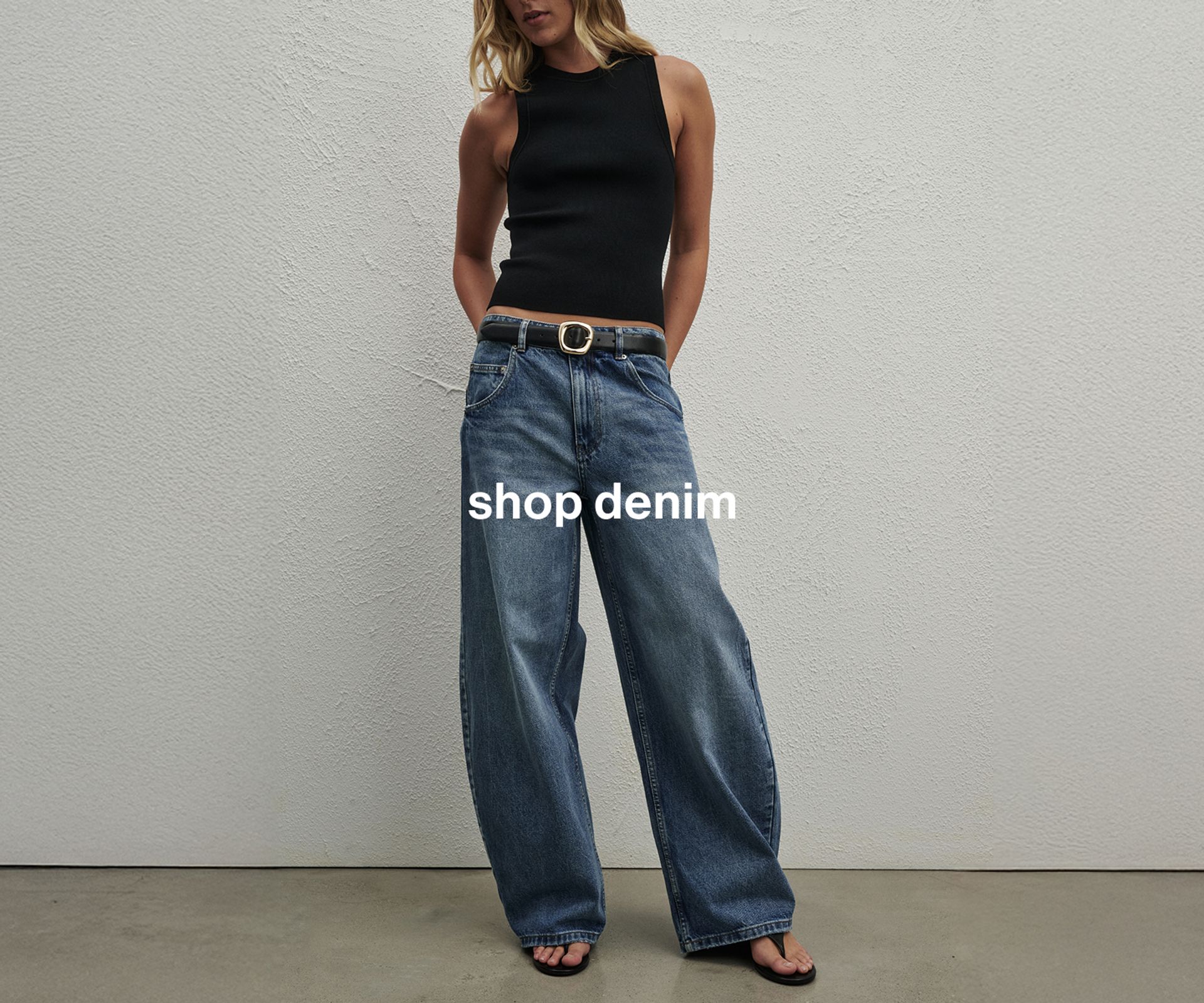 Woman wearing a black top, jeans, and a belt, standing in front of a white wall
