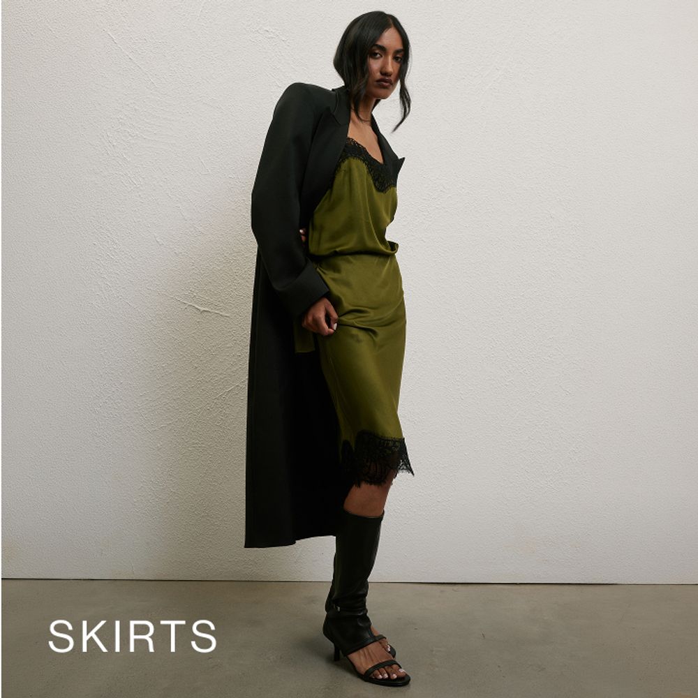 Woman wearing a long black coat, green silk top and matching skirt standing in front of a white wall with text that states "skirts"